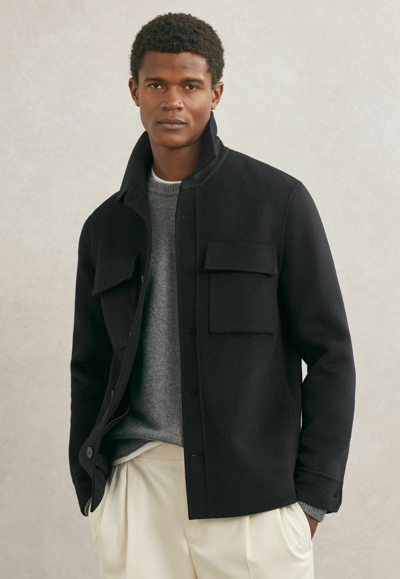 Chaqueta negra de lana con cuello alto, botones delanteros y dos bolsillos en el pecho. Llevada sobre un suéter gris y pantalones de color crema.