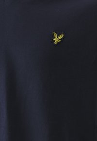 Námořnická modrá v-neck tričko vyrobené z bavlny; obsahuje malé vyšívané žluté logo orla na levé části hrudi. Hladká textura.