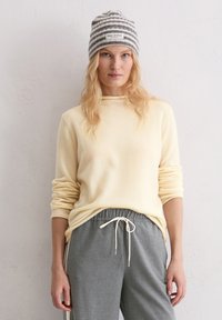 Miękki, jasnokremowy sweter z podwiniętymi rękawami w zestawieniu z szarymi spodniami dresowymi z sznurkiem. Tło jest neutralne i teksturowane.