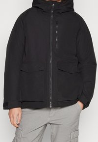 Svart pufferjacka med dragkedja framtill, stora framfickor och huva. Har slät textur och justerbara ärmslut med kardborre.