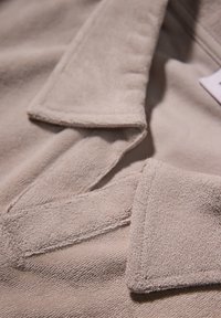 Jasno beżowa tkanina o miękkiej, teksturowej powierzchni. Posiada złożony kołnierzyk i mankiety, z widocznymi detalami stębnowania.