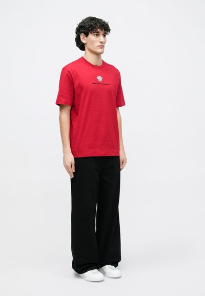 Κόκκινο βαμβακερό μπλουζάκι με στρογγυλή λαιμόκοψη και κοντά μανίκια, με γραφικό σχέδιο στο στήθος, συνδυασμένο με μαύρο παντελόνι φαρδιάς γραμμής και άσπρα παπούτσια.