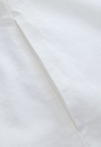 Gros plan d'un tissu blanc avec une couture visible et une texture subtile, montrant un léger pli le long de la ligne de couture.