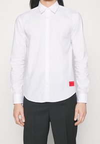 Vit långärmad skjorta i bomull med klassisk krage, som har ett rött varumärkesmärke på vänster sida nära fållen.