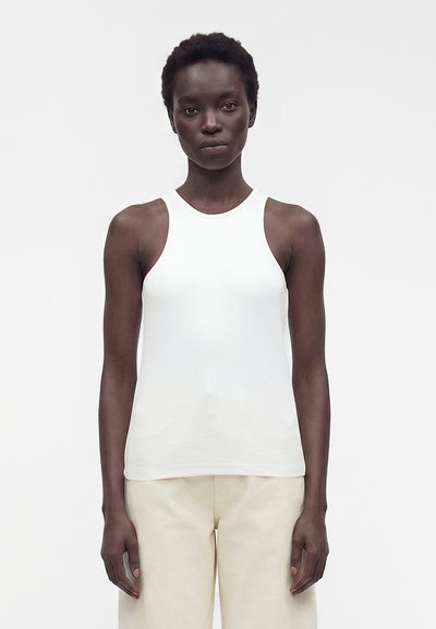 Frau mit kurzen lockigen Haaren, die ein weißes ärmelloses Top und beige Hosen trägt, steht vor einem schlichten hellen Hintergrund.