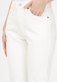 Los jeans de mezclilla blanca presentan un diseño de cintura alta, pierna recta, detalles de costura superior y un cierre de botón de metal con acentos de cobre en contraste.