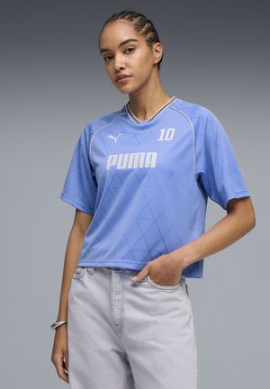 Vrouw met gevlochten haar, gekleed in een lichtblauw Puma sportshirt met nummer 10 en lichtgewassen spijkerbroek, staand met één hand in haar broekzak.