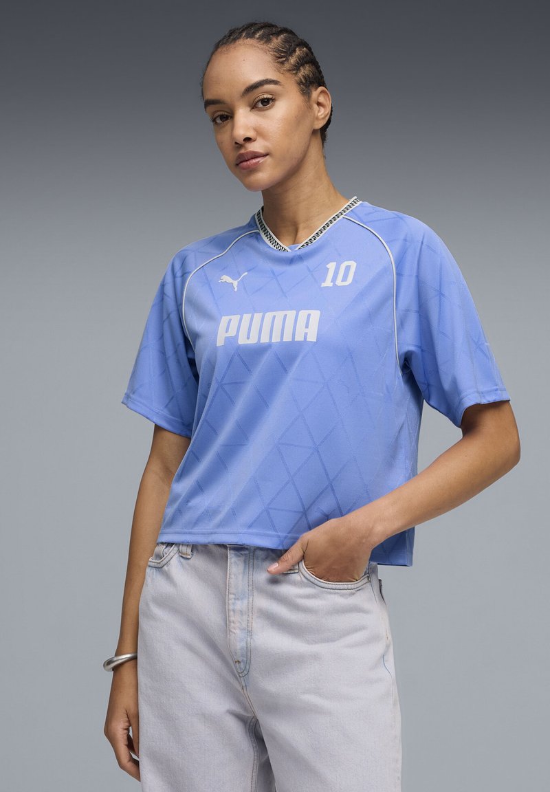 Femeie cu păr împletit purtând un tricou sport Puma albastru deschis cu numărul 10 și blugi deschiși la culoare, stând cu o mână în buzunar.