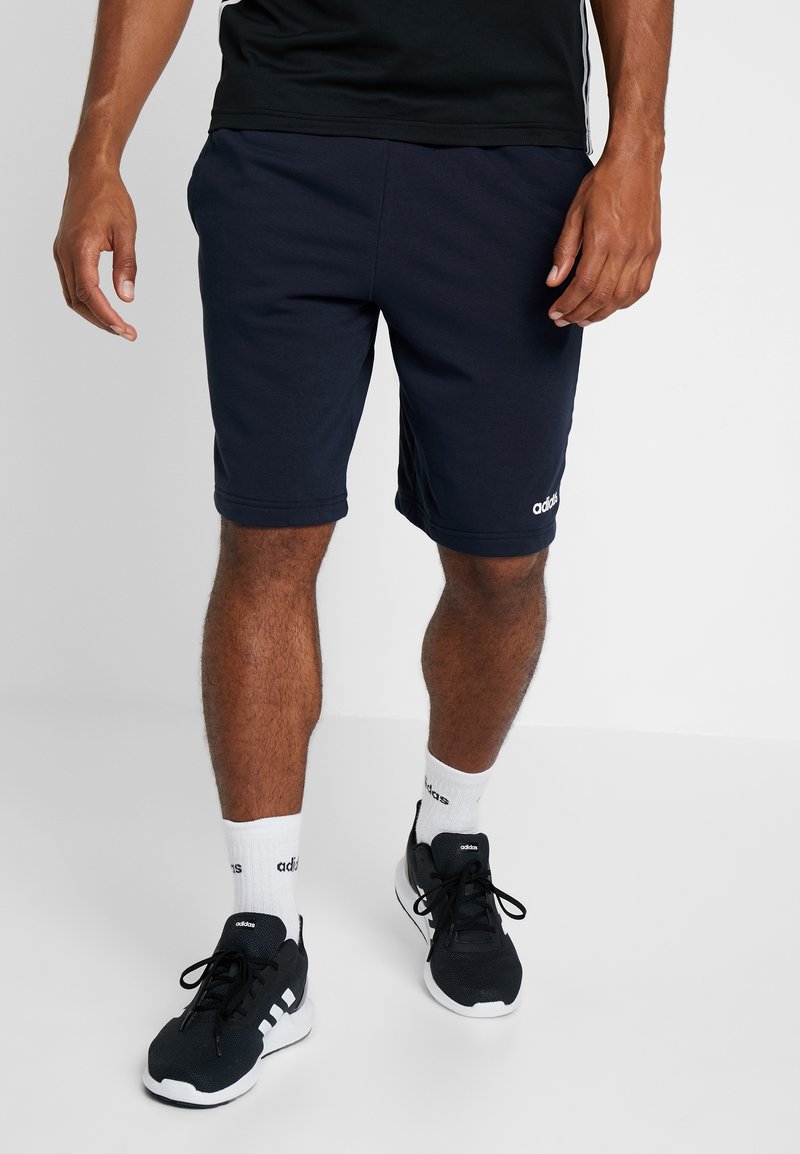 Homme portant un short noir Adidas, des baskets noires Adidas avec semelles blanches, et des chaussettes blanches Adidas, debout sur un fond clair uni.