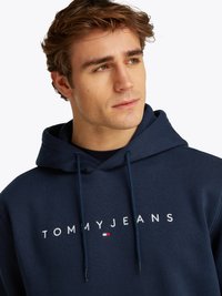 Jonge man die zijwaarts kijkt, gekleed in een marineblauwe hoodie met de tekst "TOMMY JEANS" en een klein vlaglogo op de voorkant.
