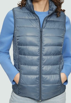 Person wearing a blue quilted sleeveless vest with hands in pockets over a long-sleeve light blue shirt, cropped at the neck and hips.