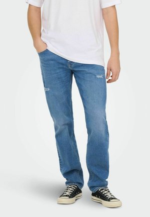 Person iført falmet blå jeans med let slid nær lommerne, sorte sneakers med hvide snørebånd og en hvid kortærmet skjorte.