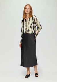Langearmige shirt in groen, roze en zwart abstracte print, gecombineerd met een zwarte satijnen midi-rok en zwarte haksandalen.