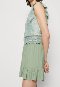 Top en dentelle florale vert clair avec des détails en feston, associé à une jupe verte froncée avec un ourlet volé. Silhouette décontractée.