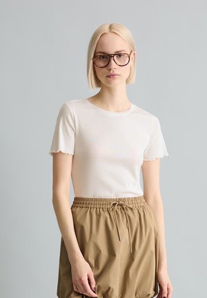 Junge Frau mit kurzen blonden Haaren, die große runde Brillen, ein weißes kurzärmeliges Oberteil und beige Hose mit Kordelzug trägt, steht vor einem grauen Hintergrund.