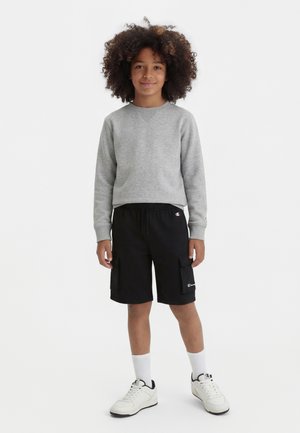 Dziecko z kręconymi włosami, ubrane w szarą bluzę z długim rękawem, czarne spodenki cargo, białe skarpetki i białe trampki, stoi i się uśmiecha.