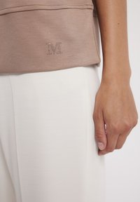 Brązowy bawełniany top z subtelnym wzorem "M" z cyrkoniami, w połączeniu z białymi teksturowanymi spodniami, które prezentują gładkie wykończenie materiału.