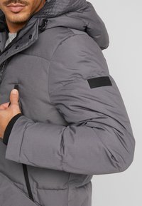 Giubbotto infilabile grigio con cappuccio, chiusura con zip frontale, tessuto strutturato e una patch nera sul maniche. Polsini a manica intera.