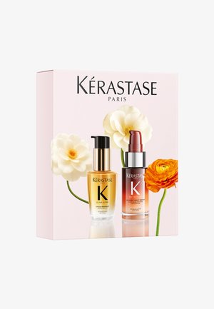 Deux bouteilles de sérum capillaire Kérastase Paris avec distributeurs pompe, sur un fond de boîte rose avec des fleurs blanches et orange.