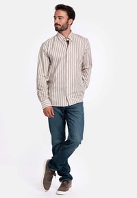 Man staat met één gebogen been, draagt een beige en wit gestreept overhemd met knopen, blauwe jeans en grijze sneakers.