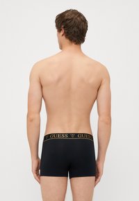 Černé boxerky s zlatým logem "GUESS" na pásku. Hladká látka, přiléhavý střih a délka do poloviny stehna. Bez vzorů nebo akcentů.