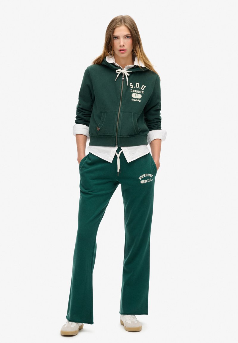 Grøn zip-up hættetrøje med frontlommer og hvid snøre, lagdelt over en hvid skjorte. Matchende grøn sweatpants med logo detaljer.
