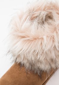 Slipper de ante marrón con un puño de pelaje sintético esponjoso de color rosa claro y acentos de pelo más oscuro, que enfatizan la textura y la apariencia suave y acogedora.