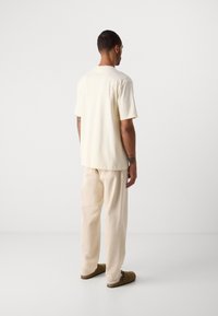 Uomo di spalle che indossa una t-shirt oversize color bianco sporco, pantaloni crema e scarpe slip-on marroni su uno sfondo bianco uniforme.