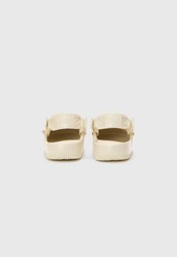 Beige Nike čības ar gludu tekstūru, atvērtu aizmuguri, regulējamu siksnu un rievotu zoli. Zīmols iegravēts uz siksnas.
