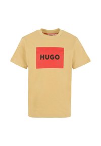 T-shirt beige à manches courtes avec un logo rectangulaire rouge affichant le mot "HUGO" en lettres noires en gras à l'avant.