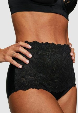 Vrouw draagt hoog getailleerd zwart kanten ondergoed en een zwarte bh, handen op de heupen, met aandacht voor het kanten detail van het ondergoed.