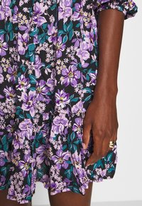 Blumenkleid mit lila und türkisfarbenen Blumen auf einem schwarzen Hintergrund. Mit gerafftem Saum und gesammelten Ärmeln. Die Hand des Models ist sichtbar und trägt Ringe.