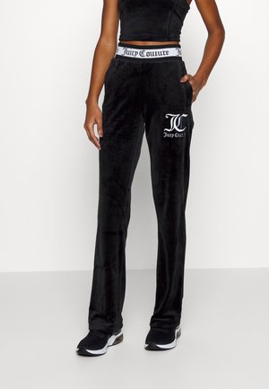 Osoba nosząca czarne welurowe spodnie Juicy Couture z białym logo na udzie i markowanym elastycznym pasem, zestawione z czarnymi trampkami.