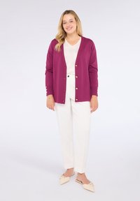 Magenta svetr s perlovými knoflíky na zapínání, nošený přes světlo béžový top a bílé kalhoty. Svetr má dlouhé rukávy a volný střih.