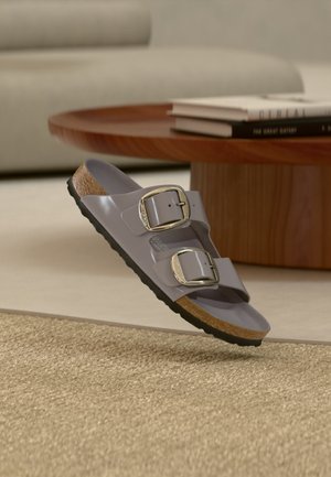 Ένα γκρι σανδάλι με δύο μεγάλες αγκράφες αιωρείται πάνω από μια υφασμάτινη μπεζ μοκέτα, δίπλα σε ένα στρογγυλό ξύλινο τραπεζάκι με στοιβαγμένα βιβλία.