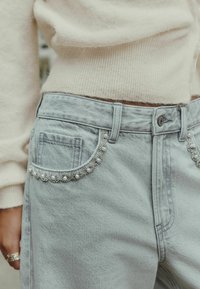 Világosszürke farmernadrág, szoros szabással. Dekoratív gyöngy részletekkel a hátsó zseb körül és normál gombzárral rendelkezik.