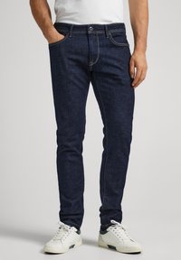 Mörkblå denimjeans med slim fit, femfickorsdesign, synlig sömnad och knapp- och dragkedjestängning. Bärs med vita sneakers.