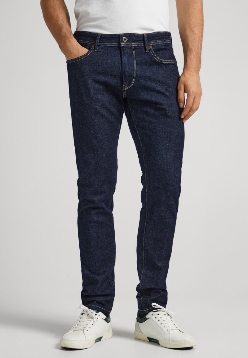 Mörkblå denimjeans med slim fit, femfickorsdesign, synlig sömnad och knapp- och dragkedjestängning. Bärs med vita sneakers.