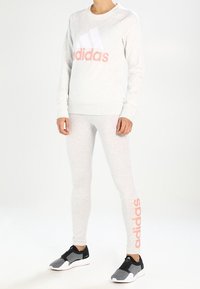 Jasnoszary sweatshirt z białym i różowym logo "adidas", w zestawie z pasującymi jasnoszarymi legginsami i czarnymi butami sportowymi z białymi podeszwami.