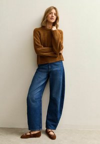 Suéter marrón tejido, vaqueros azules de pierna ancha y zapatos planos marrones con detalles de metal. El conjunto presenta un ajuste casual y texturas suaves.