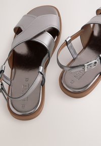 Sandales métalliques argentées avec un design à bretelles croisées, bride de cheville réglable, semelle intérieure rembourrée et semelle en caoutchouc marron. Texture lisse.