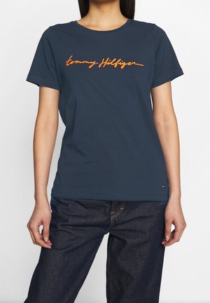 Žena nosící tmavě modré tričko Tommy Hilfiger s oranžovým logem a tmavě modré džíny, stojící na jednobarevném světlém pozadí.