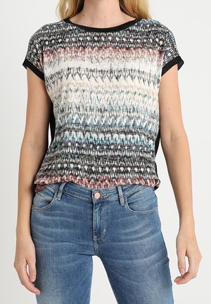 Femme portant un haut à manches courtes à motifs multicolores avec des bordures noires et un jean bleu, debout devant un fond uni.