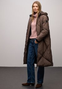 Brązowy pikowany długi płaszcz z kapturem, wzór w romby, w połączeniu z jasnoróżowym golfem i niebieskimi dżinsami. Brązowe botki za kostkę.