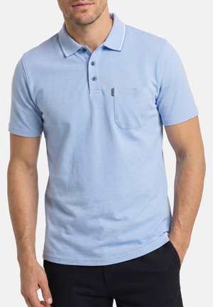 Mann trägt hellblaues kurzärmeliges Poloshirt mit Kragen und Brusttasche, Hände entspannt, kombiniert mit dunkler Hose.