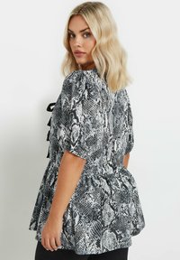 Blusa de estampado de serpiente en blanco y negro con mangas cortas acampanadas, dobladillo peplum y adornos de cinta negra en la parte posterior. Textura de tela suave.