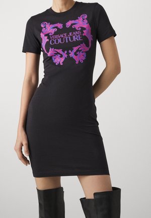 Musta vartalonmyötäinen lyhythihainen mekko, jossa vaaleanpunainen ja violetti kukkakuvio sekä "Versace Jeans Couture" -teksti, yhdistetty mustiin polvipitkiin saappaisiin.