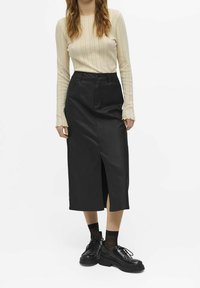 Jupe crayon en faux cuir noir avec une fente frontale, associée à un haut à manches longues côtelé beige clair ajusté et des chaussures noires épaisses.