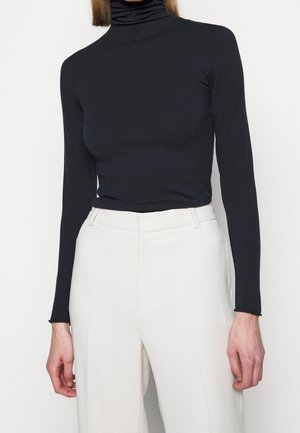 Personne portant un col roulé noir ajusté et un pantalon blanc taille haute, debout devant un fond uni.