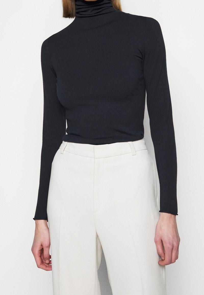 Personne portant un col roulé noir ajusté et un pantalon blanc taille haute, debout devant un fond uni.